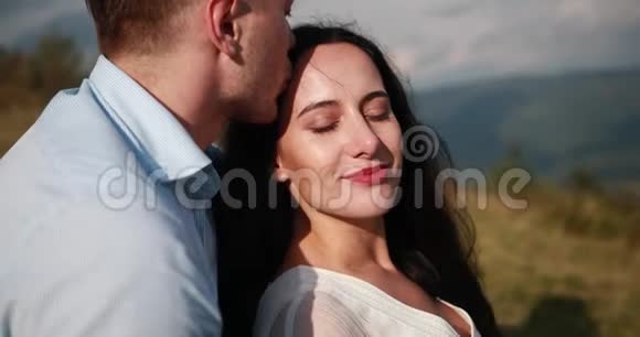 男人温柔地拥抱黑发女人亲吻她坐在山顶上的前额视频的预览图