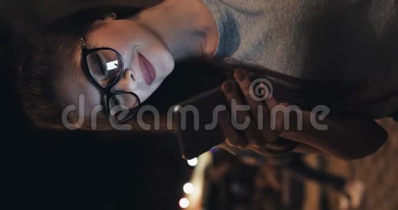 戴眼镜的迷人年轻女子坐在夜市街道的长凳上用智能手机对视频的预览图