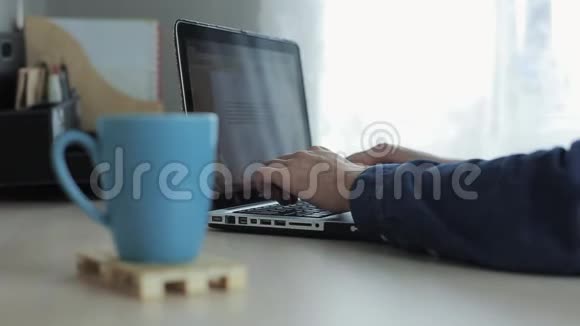 在背景上的笔记本电脑上用男性的手打着字关上一杯茶或咖啡在办公室工作的家庭自由职业者视频的预览图