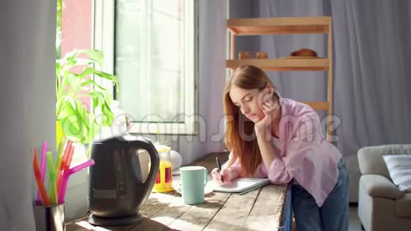 年轻女子站在厨房的木桌旁喝茶做笔记视频的预览图
