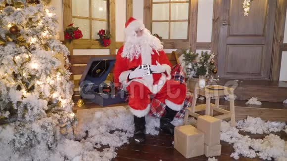 圣诞老人正包着礼物或礼物坐在屋外的圣诞树间喝着牛奶听着视频的预览图