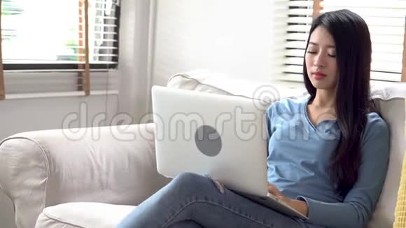 美丽的亚洲年轻女性坐在沙发上卧室里放着笔记本电脑和手机视频的预览图