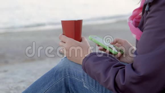 4K那个女人坐在海滩上喝着一杯热饮料用她的手机从后面近距离拍摄视频的预览图