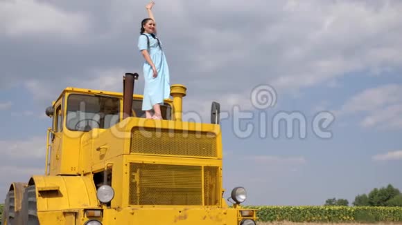 年轻女子站在黄色的拖拉机上在蓝蓝的天空中缓慢地向你挥手视频的预览图