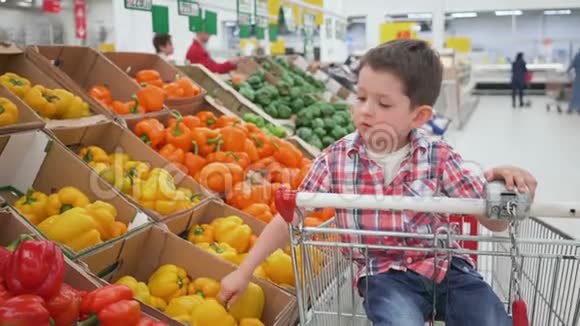 超市里的小男孩坐在手推车里闻着黄色的保加利亚胡椒商店购物新鲜产品视频的预览图