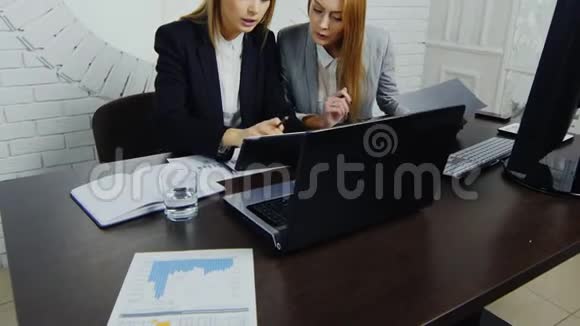 两位穿着严格西装的年轻商务女士在办公室工作坐在桌子旁看着平板电脑视频的预览图
