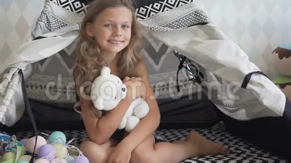 可爱的小女孩抱着她的玩具熊坐在帐篷里的地板上看着相机视频的预览图