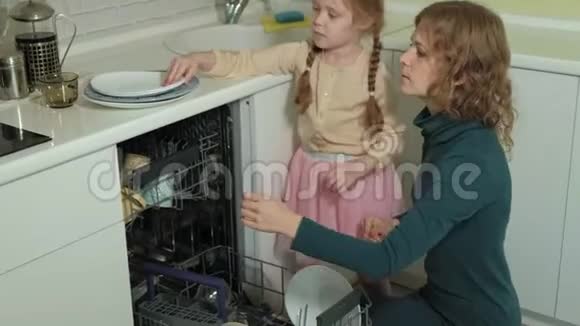 可爱的金发女婴穿着粉红色的裙子在明亮的厨房里帮助妈妈从洗碗机里取出盘子和杯子视频的预览图