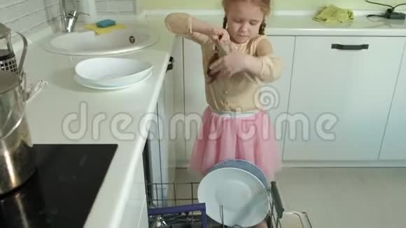 可爱的金发女孩穿着粉红色的裙子在明亮的厨房帮助妈妈在洗碗机里装盘子杯子视频的预览图