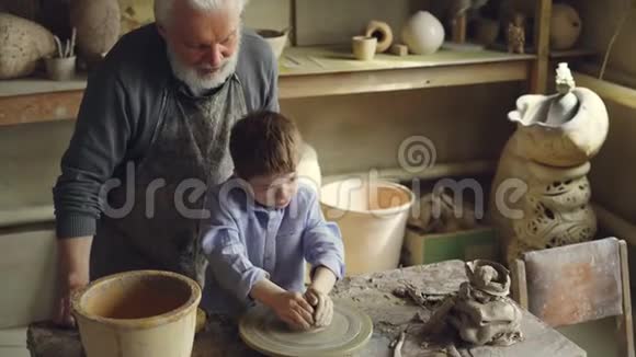年轻可爱的男孩正在投掷轮子上做泥人而他的祖父有经验的雕刻家站在附近视频的预览图