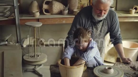 可爱的男孩在碗里润湿双手在投掷轮上浇灌粘土人物而他的祖父有雕刻经验视频的预览图