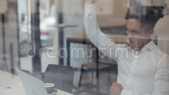 快乐的年轻商人坐在一张桌子上在一家咖啡馆里用笔记本电脑工作赢家看手提电脑显示是的手势视频的预览图
