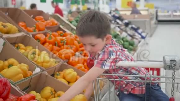 超市里的小男孩闻着坐在手推车里的黄色保加利亚辣椒商店购物新鲜产品视频的预览图