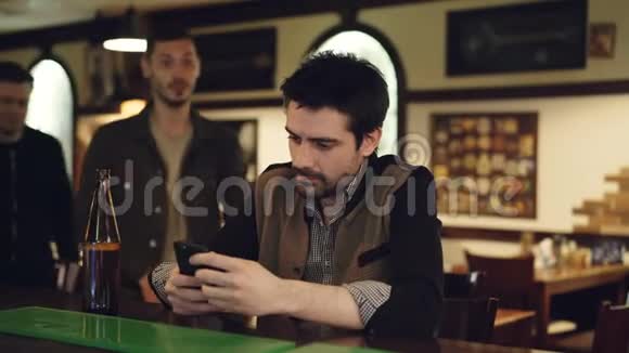 年轻的帅哥在酒吧柜台使用智能手机时他的男性朋友来了看着屏幕讨论着视频的预览图