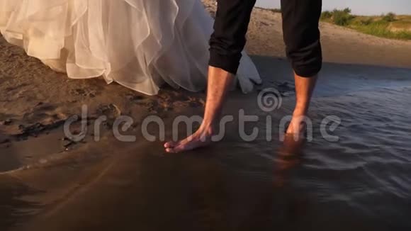 可爱的夫妇在夏天的海滩上散步新娘穿着白色礼服新郎赤脚在水面上行走视频的预览图