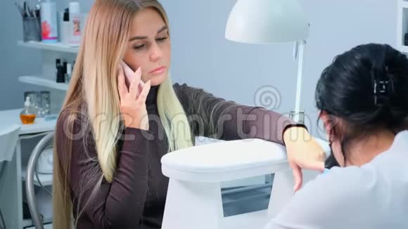 美甲师为美容院的女人做美甲一个金发女人在修指甲时打电话视频的预览图