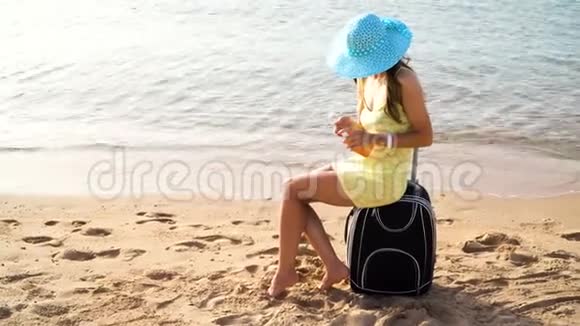夏天旅行时戴着一顶帽子手里拿着一罐防晒霜的漂亮女人坐在一个箱子上背着大海视频的预览图