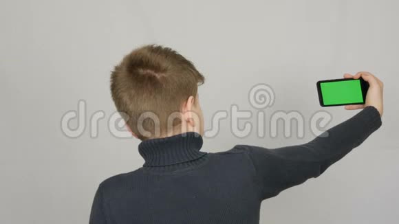 一个十几岁的男孩背对着相机站着手里拿着一部黑色智能手机上面有一个白色的绿色屏幕视频的预览图