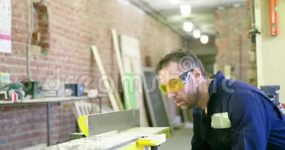 一个穿着工作服和安全眼镜的人把机器上的锯末吹掉木匠车间工作视频的预览图