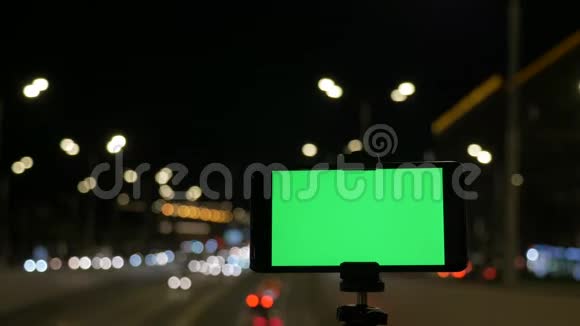 带有绿色屏幕的智能手机安装在三脚架上在汽车红绿灯的背景下晚上视频的预览图