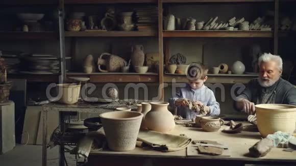 熟练的孩子正在帮助他的高级祖父专业陶工捏粘土手工陶瓷人物家庭视频的预览图