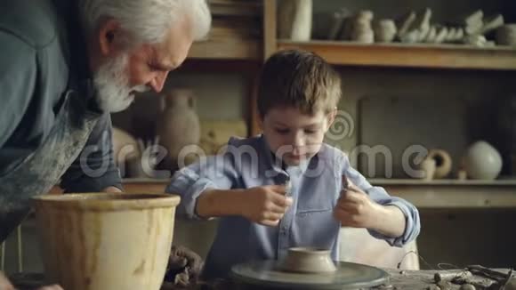 小男孩陶器学家正在制作投掷轮上的陶器而他有经验的爷爷正在和他说话视频的预览图