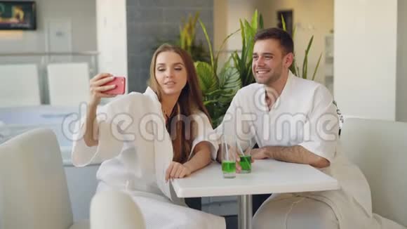 漂亮的女孩和她可爱的男朋友在水疗中心放松时用智能手机用鸡尾酒杯自拍视频的预览图