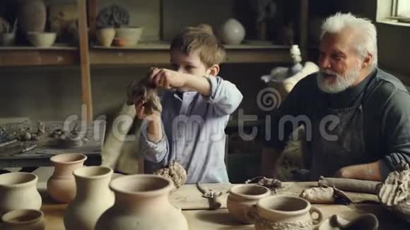专业的老陶工正在看他的孙子在他的陶器工作室里玩粘土泥里的创意男孩视频的预览图