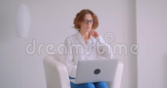 戴眼镜的白种人成年女商人用笔记本电脑坐在扶手椅上的特写肖像视频的预览图