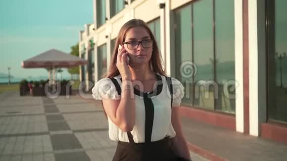 穿着白色衬衫和眼镜的女商人在一座现代化办公楼附近的街道上散步在电话里交谈视频的预览图