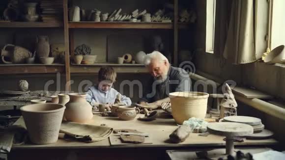 有经验的工匠教他的孙子陶器艺术并为陶瓷人物制作粘土卷而好奇的男孩视频的预览图