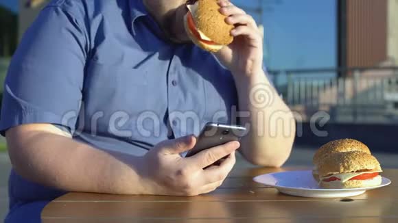 懒惰的年轻人吃不健康的汉堡和滚动智能手机应用程序视频的预览图