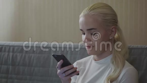 家中客厅的沙发上坐着一个暖色调的金发女郎用智能手机在线识别声音视频的预览图