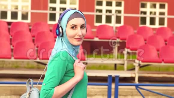漂亮的穆斯林女孩戴着头巾在体育场周围走来走去对着镜头微笑画像视频的预览图