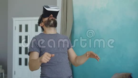 穿着休闲服装的大胡子男子正在使用虚拟现实眼镜站在他的公寓里做手势现代视频的预览图