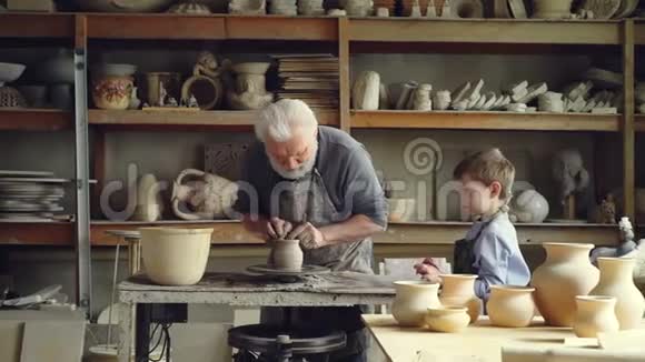 资深陶工正在纺抛轮成型粘土而他的小孙子正在观看和学习分享视频的预览图
