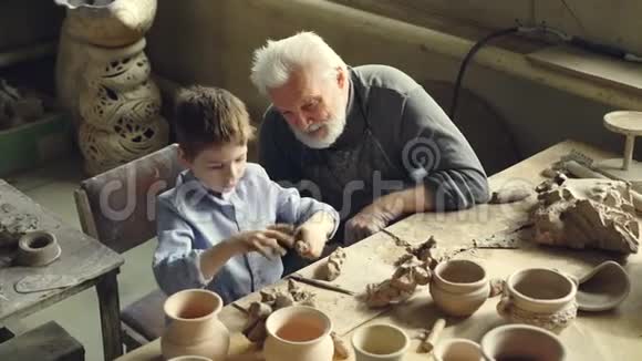 好奇的小男孩一边捏着黏土一边和爷爷的职业陶工说话一边坐在桌子上视频的预览图