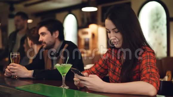 年轻的女学生正在使用智能手机坐在高档酒吧和鸡尾酒她摸着屏幕微笑着现代视频的预览图