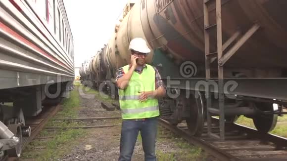 一个戴眼镜的人穿着头盔和制服检查火车走在火车站上打电话视频的预览图