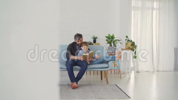 长胡子的男人正在看书给他的儿子看插图同时在光线公寓的沙发上放松联合视频的预览图