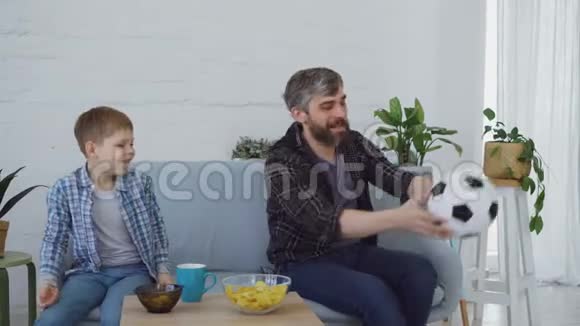 快乐可爱的父亲和他的小儿子一起在小公寓里踢足球和吃零食视频的预览图