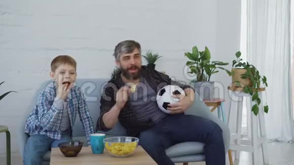 专心致志的爸爸和小儿子正在家里看电视上的足球赛欢呼雀跃庆祝胜利视频的预览图
