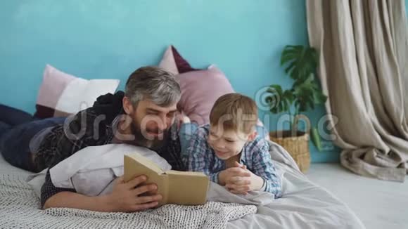 关爱的父亲正在给他的孩子读有趣的书而男孩正在笑着和他的父母说话全尺寸床明亮视频的预览图