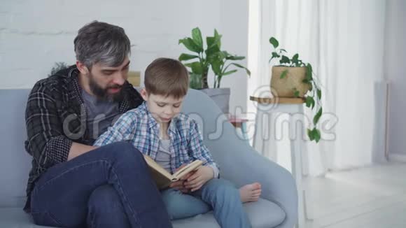 可爱的父亲正在教他的孩子认真的学龄前儿童一边坐在沙发上看书绿色植物视频的预览图