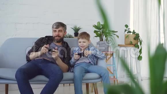 父亲和他的孩子正在家里的沙发上玩电子游戏按下操纵杆上的按钮和交谈快乐视频的预览图