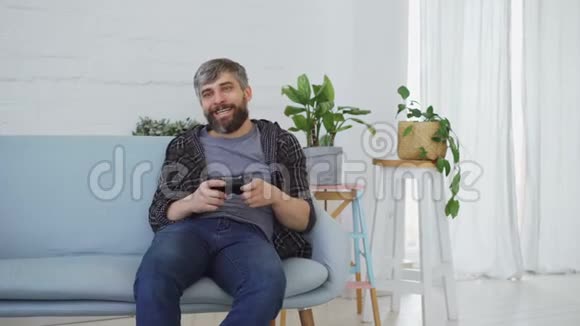 英俊的胡须男穿着休闲服正坐在家里的沙发上玩电子游戏现代科技快乐视频的预览图
