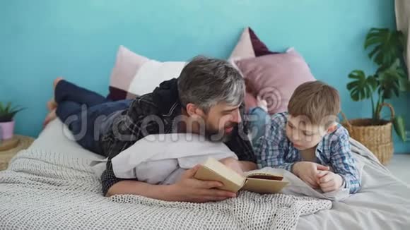 长胡子的男人正在给他的小儿子看书他躺在舒适的卧室里躺在床上团结家庭消磨闲暇时间视频的预览图
