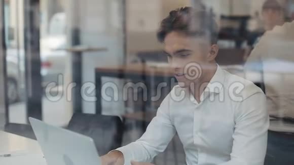 快乐的年轻商人坐在一张桌子上在一家咖啡馆里用笔记本电脑工作赢家看手提电脑显示是的手势视频的预览图