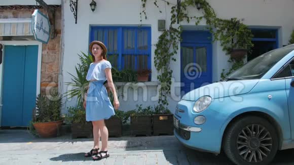 年轻的红发美女穿着蓝色连衣裙在复古车前在街上蹦蹦跳跳视频的预览图