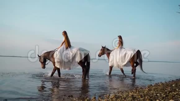 两个穿着白色长裙的漂亮女孩骑马骑马的人在日落时沿着海岸漫步视频的预览图